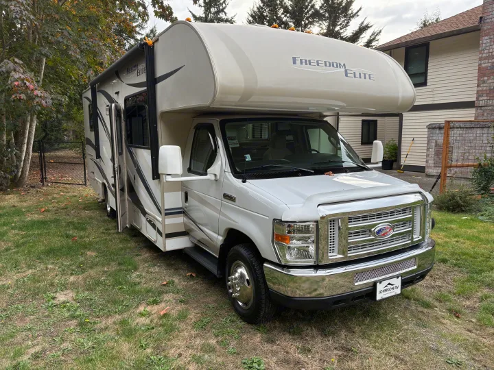 This white Freedom Elite Class C **rv rental**, built on a Ford chassis, is parked on a grassy lawn next to a house.. Thor Freedom Elite 2014