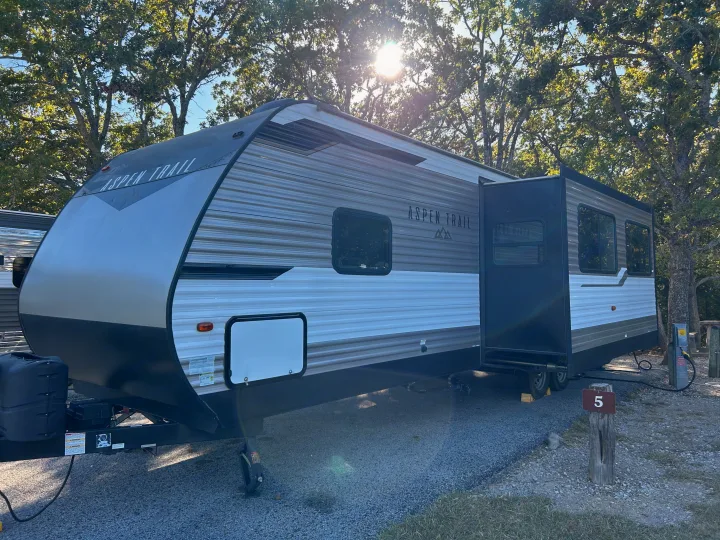 RV rental "Aspen Trail" travel trailer parked at a sunny campsite (site 5) among green trees.. Dutchmen 3120BHS 2021