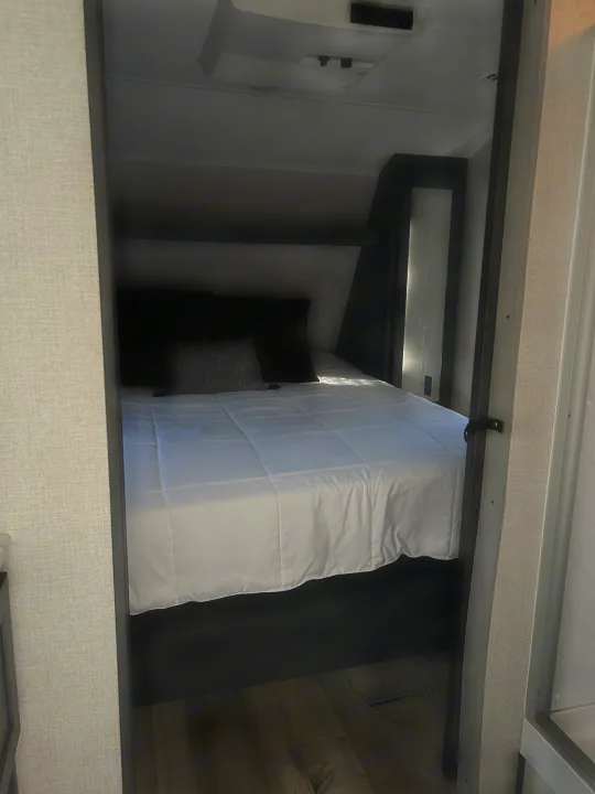 Step into a cozy, compact RV rental bedroom with a neatly made bed, white bedding, dark pillows, and wood flooring.. Dutchmen 3120BHS 2021