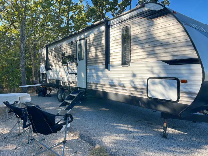 Experience outdoor adventures in this Aspen Trail RV rental. Parked at a gravel campsite with trees, chairs, and a picnic table on a sunny day.. Dutchmen 3120BHS 2021
