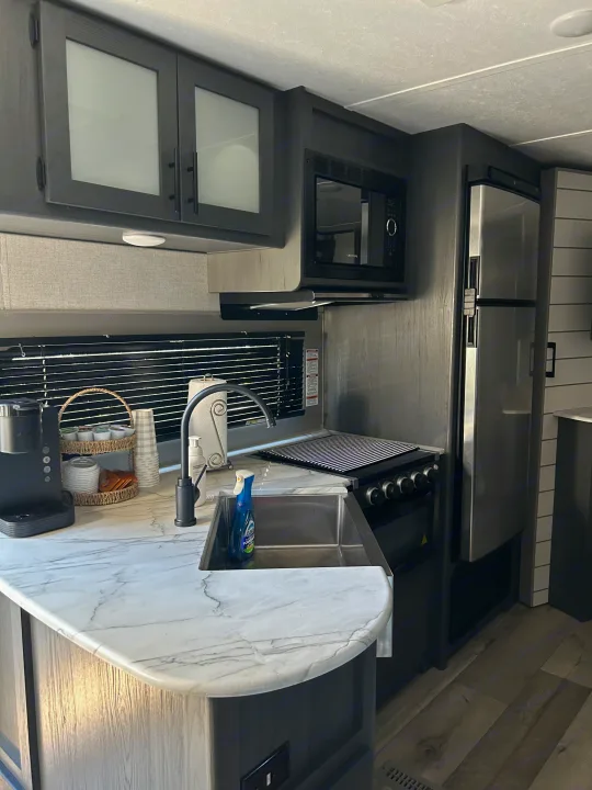 Stylish RV rental kitchen featuring dark cabinetry, marbled counters, coffee station, sink, and stainless appliances.. Dutchmen 3120BHS 2021