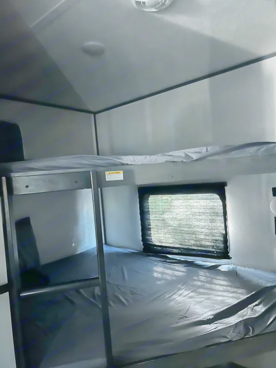 RV rental bunk beds, dimly lit with grey sheets. A caution sticker is visible, and a window shows outdoor greenery through blinds.. Dutchmen 3120BHS 2021