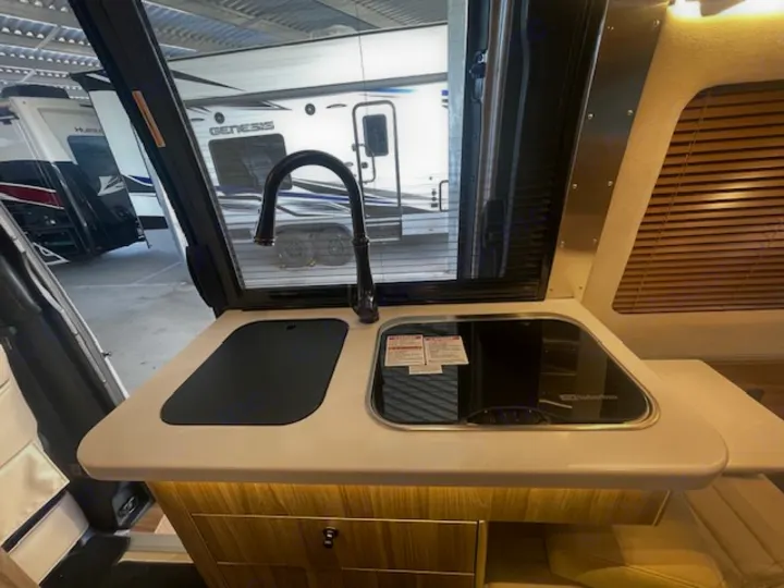 Explore this RV rental's compact kitchen: modern sink, dark faucet, sleek glass cooktop, and wooden cabinets. Ideal for travel.. Airstream Interstate 2023