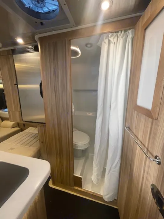 This compact RV rental interior features a wet bath with toilet & shower curtain, plus partial kitchen and sleeping area.. Airstream Interstate 2023