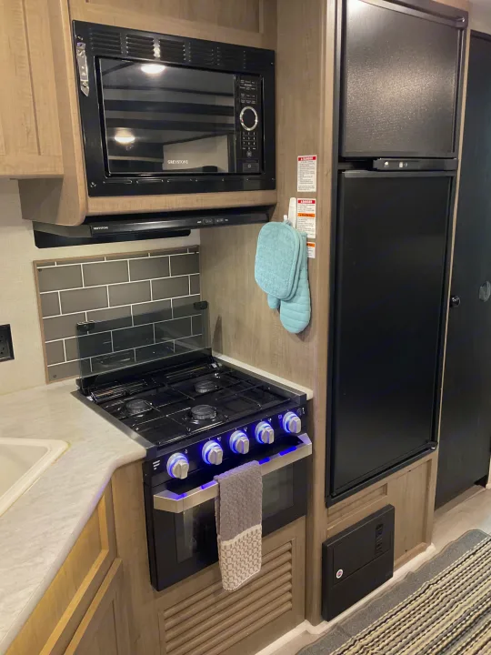 RV rental's modern kitchen: microwave, gas stove/oven with blue lights, fridge, sink, and tiled backsplash.. Keystone RV Hideout 2021