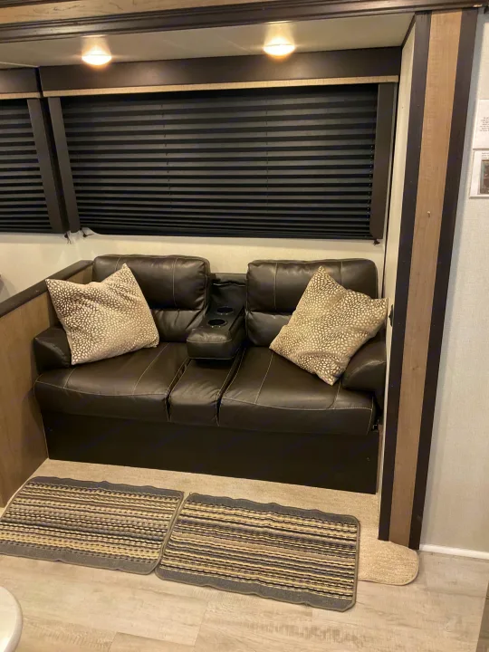 Comfortable RV rental interior featuring a leather loveseat with cup holders, patterned pillows, black blinds, and striped rugs.. Keystone RV Hideout 2021