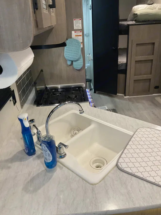 Compact rv rental kitchen: double sink, stove, cleaning supplies, and bunk beds visible, showcasing efficient design.. Keystone RV Hideout 2021