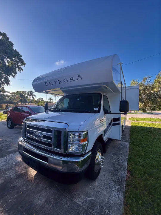 White Entegra rv rental, a Class C motorhome, on a sunny day. Parked on a driveway with trees and a red car nearby.. Entegra Ford Transit 2025