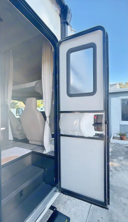 Open door of an RV rental, revealing the interior with steps, a driver's seat, and an overhead bunk. Ready for your next trip!. Entegra Ford Transit 2025