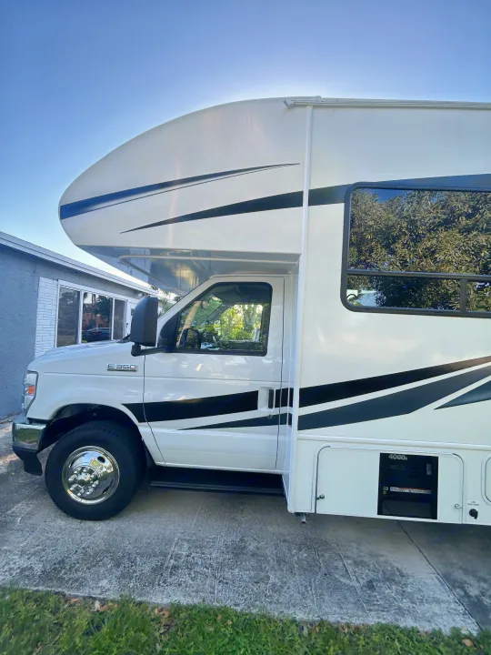 This white Class C RV rental, with sleek black stripes, is parked on concrete, ready for adventure. Generator compartment visible.. Entegra Ford Transit 2025