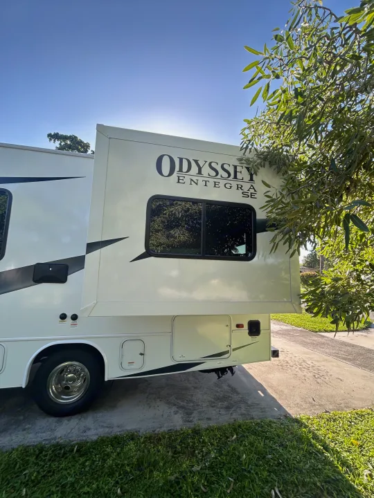 Odyssey Entegra SE RV rental parked with its slide-out extended on a sunny day, partially framed by lush green tree branches.. Entegra Ford Transit 2025