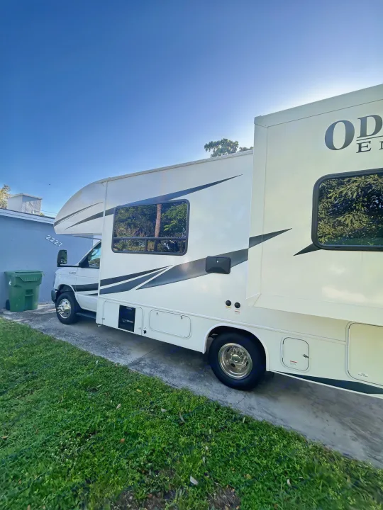 White RV rental (likely Odyssey E350) parked on a driveway under a clear sky, showing its side, windows, and storage.. Entegra Ford Transit 2025