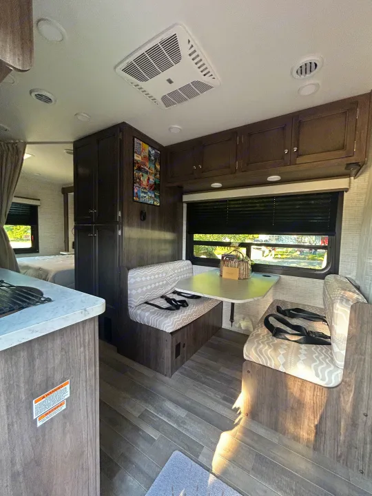 Inside an RV rental: Dinette with seatbelts, compact kitchen, and a glimpse of the bedroom, designed for travel convenience.. Entegra Ford Transit 2025