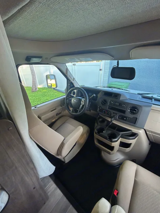 Inside an RV rental: driver's area with dashboard, steering wheel, light-colored seats, and outdoor view through windshield.. Entegra Ford Transit 2025