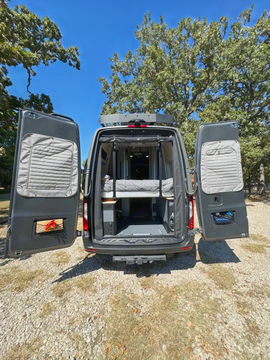 Spacious rv rental van ready for adventure. Open doors reveal bed & gear, parked on gravel under sunny skies.. Winnebago Revel 2025