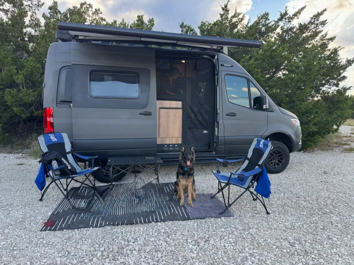 A gray camper van RV rental parked on gravel with an outdoor setup, including chairs, a table, and a long-haired German Shepherd dog.. Winnebago Revel 2025