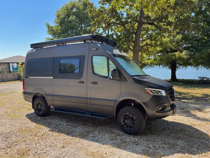 This gray custom Sprinter RV rental, with off-road tires and a roof rack, is parked by a scenic lake, perfect for adventure.. Winnebago Revel 2025