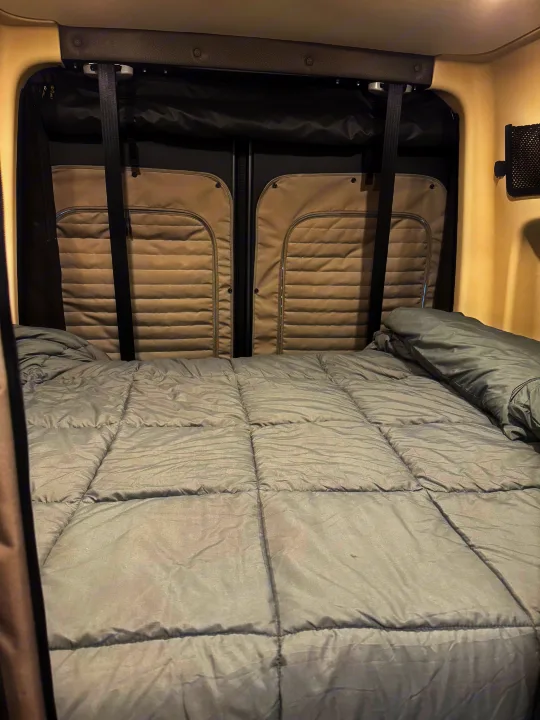 Comfortable sleeping area in an rv rental, featuring a bed with a grey comforter and padded rear door panels.. Winnebago Revel 2025