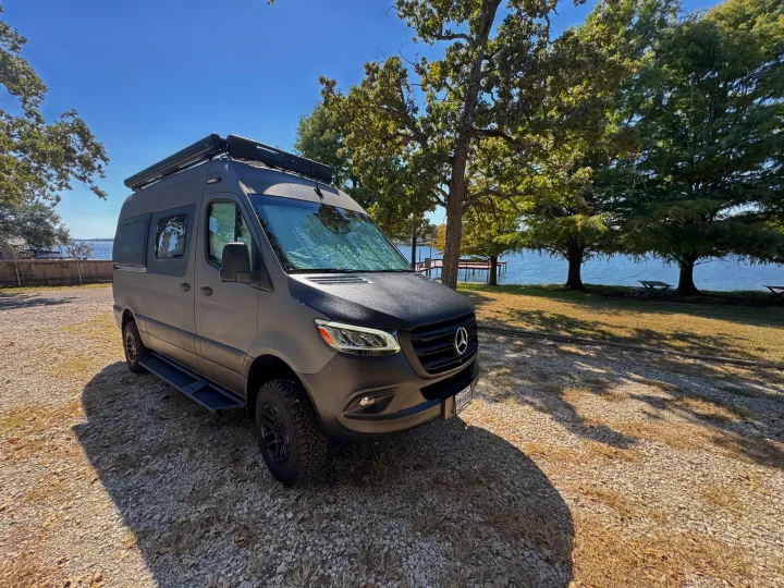 Black Mercedes Sprinter RV rental parked on a gravel path by a scenic lake and trees, perfect for a van life adventure.. Winnebago Revel 2025