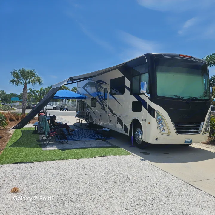 Enjoying a sunny day at an RV rental campsite. A large motorhome parked with awning extended, people relaxing outside.. Thor Windsport 2023