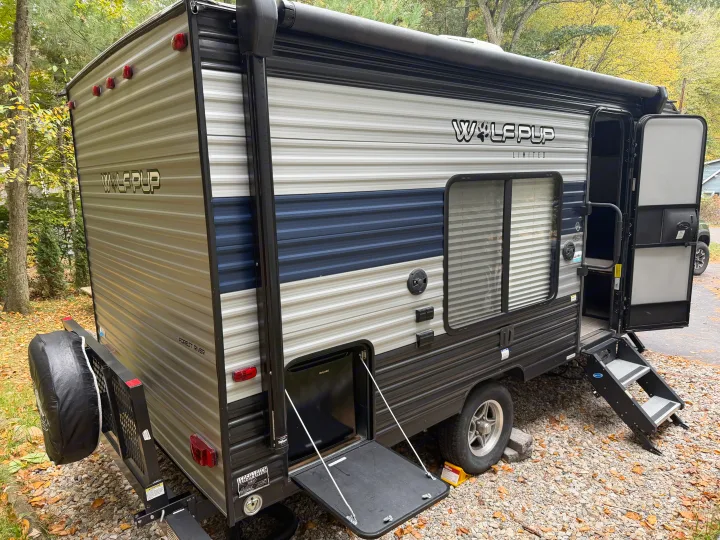 This gray and blue Wolf Pup Limited rv rental travel trailer, with open door, steps, and outdoor kitchen, sits on a gravel campsite.. Forest River Cherokee Wolf Pup 2021