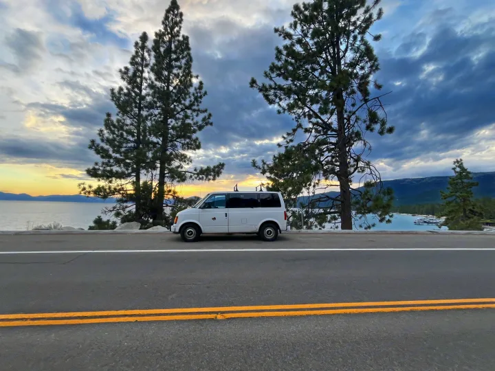 White RV rental parked roadside, overlooking a tranquil lake with sunset hues, tall pines, and distant mountains.. Chevrolet Astro 1992