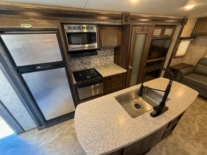 Modern RV rental kitchen with stainless appliances, an island sink, stove, and fridge. Dark wood and herringbone backsplash.. Highland Ridge RV Light 2018