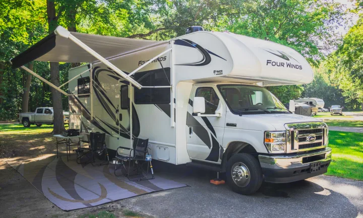 White Class C rv rental at a sunny campsite with an extended awning, outdoor rug, and chairs, ready for adventure.. Thor Four Winds 2021