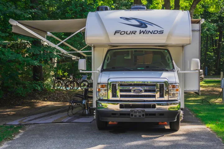 Front view of an RV rental: A Four Winds Ford motorhome with an extended awning, parked at a wooded campsite with bicycles.. Thor Four Winds 2021