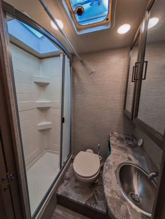 Compact RV rental bathroom featuring a glass-enclosed shower, toilet, vanity with sink, and mirror cabinet for efficient travel living.. Thor Four Winds 2021