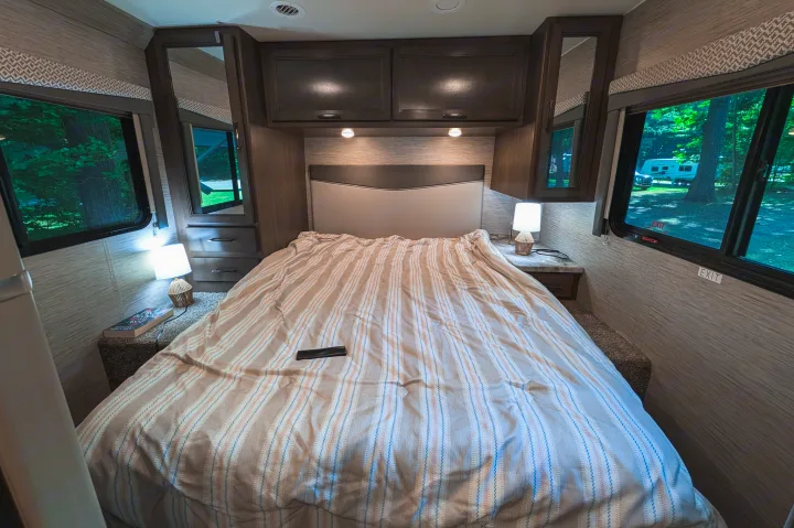 This RV rental bedroom features a comfortable striped bed, overhead storage, and windows with a view of a campsite.. Thor Four Winds 2021