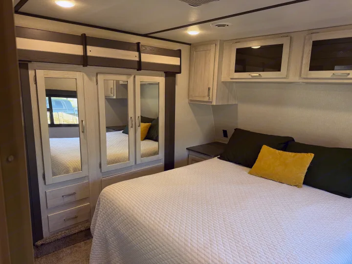 RV rental bedroom: white quilted bed with green & yellow pillows, mirrored wardrobe, and light wood overhead storage.. Keystone RV Sprinter Campfire 2021