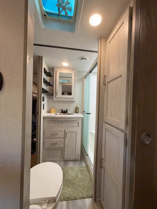 Modern RV rental bathroom: vanity, shower, toilet, and ample storage, featuring light wood finishes and a ceiling fan.. Keystone RV Sprinter Campfire 2021