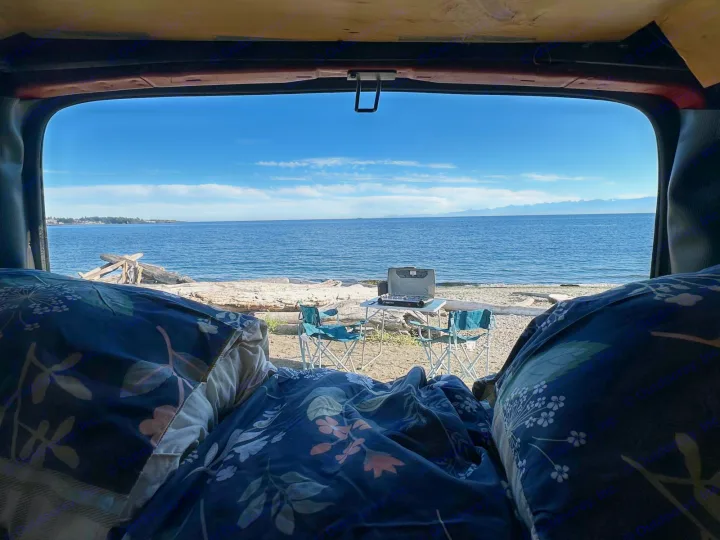 Cozy rv rental view: Floral bedding inside, looking out at a serene beach with driftwood, a portable stove, and blue ocean.. Gmc Savanna 2003