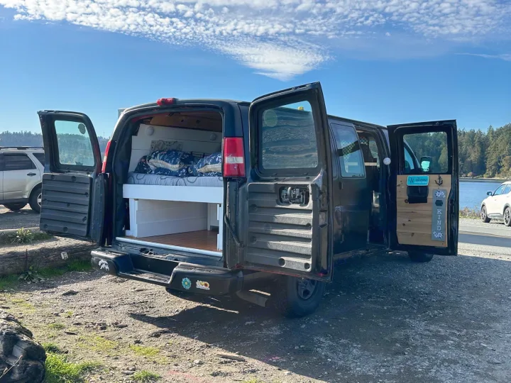 This black converted van, ready as an RV rental, displays its bed and custom wooden interior at a scenic waterside campsite under a blue sky.. Gmc Savanna 2003