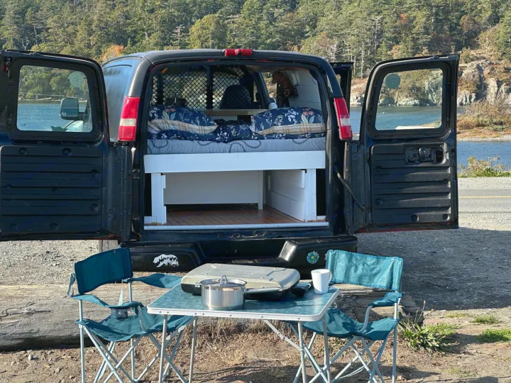 Scenic rv rental camper van with open back doors, comfy bed, and outdoor camping setup by the water. Perfect for a road trip.. Gmc Savanna 2003