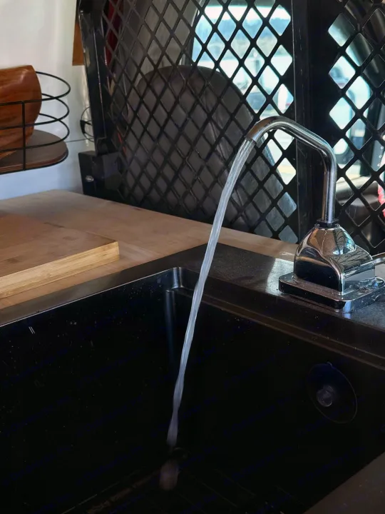 Water flows from a chrome faucet into a black sink, set in an RV rental's kitchen with a wooden counter and mesh grid.. Gmc Savanna 2003
