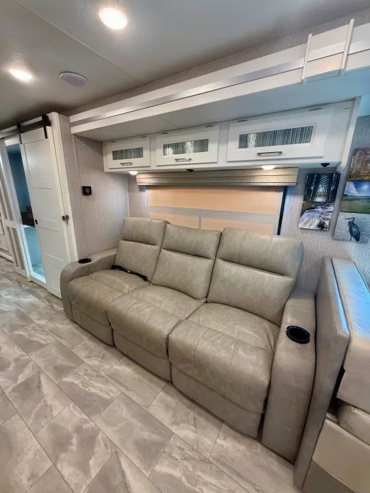 Modern rv rental living area with a plush gray reclining sofa, sleek overhead cabinets, and stylish patterned flooring. Bright and inviting.. Coachmen Encore 2022