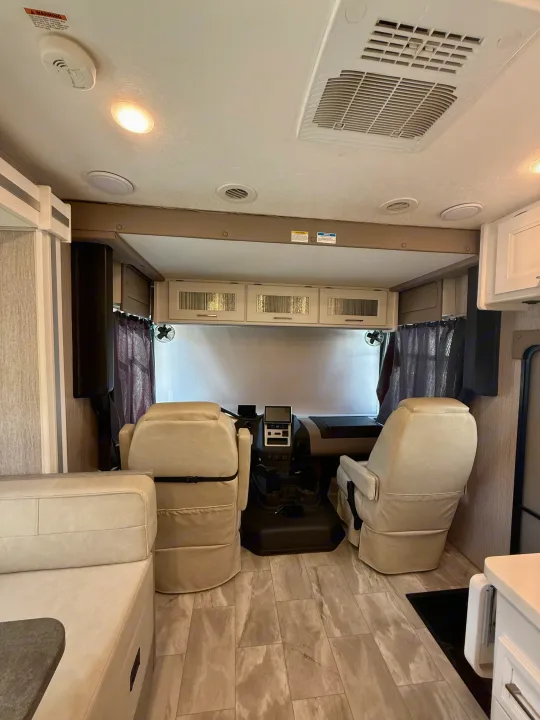 Modern RV rental interior: cockpit with tan seats, dashboard, overhead cabinets, and front window blinds and fans.. Coachmen Encore 2022