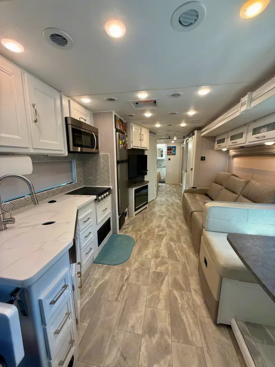 Modern rv rental interior showcasing a bright kitchen with white cabinets, comfortable living room with a grey sofa, and tile flooring.. Coachmen Encore 2022