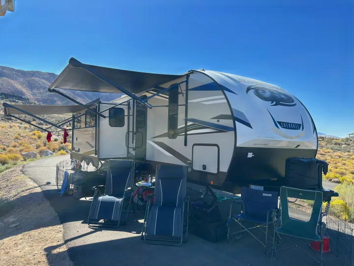 RV rental travel trailer campsite. White & grey camper with extended awnings, chairs, & pet enclosure under a clear sky, mountains in back.. Forest River Cherokee Alpha Wolf 2021