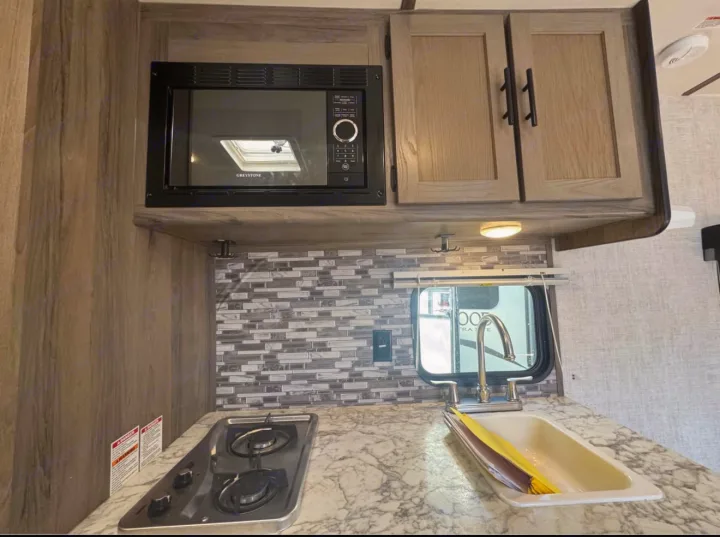 Explore this RV rental kitchen featuring a microwave, stovetop, sink, and cabinets – all ready for your journey.. Gulf Stream Kingsport Super Lite 2021