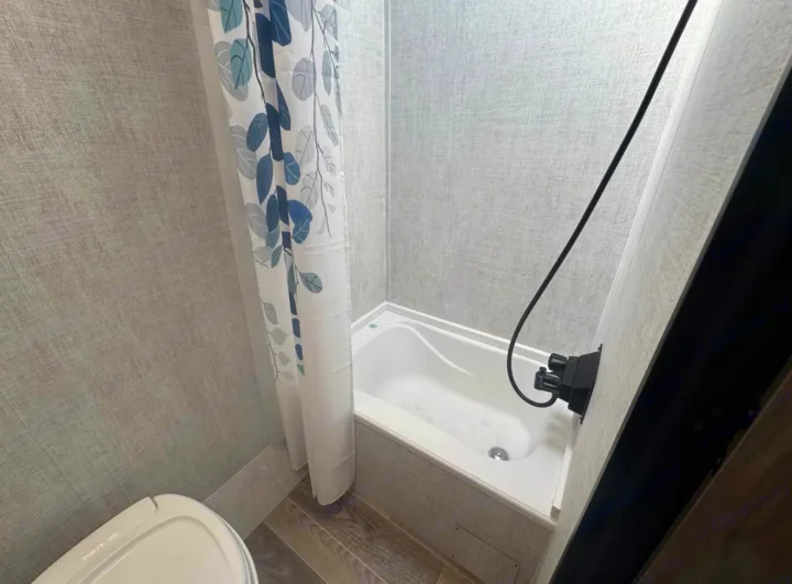 Clean rv rental bathroom with a compact white tub, black shower fixture, and a white curtain with blue and green leaf print.. Gulf Stream Kingsport Super Lite 2021