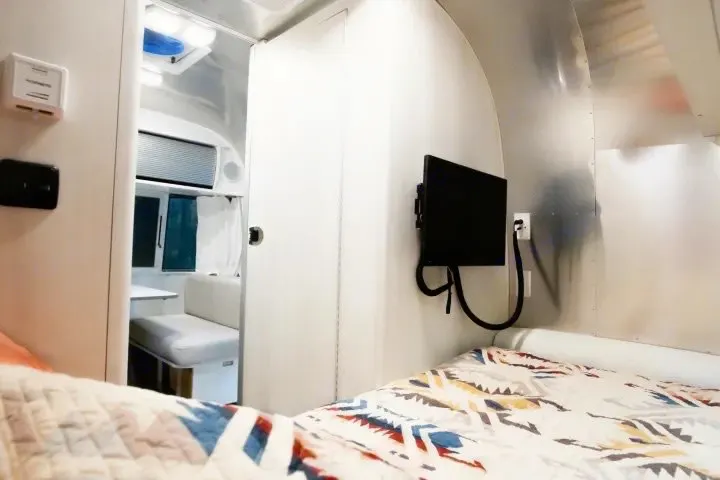 Modern rv rental interior with a cozy bed, wall-mounted TV, and view into the dinette. Sleek travel trailer design.. Airstream Bambi 2022
