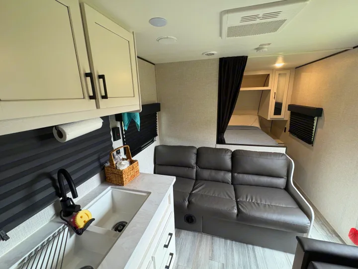 This RV rental features a compact kitchen with white cabinets, dark sofa, and a private bed area, ideal for travel.. Jayco Jay Flight SLX 2024