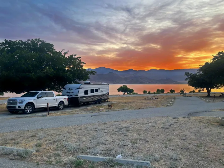 A white pickup truck tows a Jayco travel trailer at a lakeside campsite with a vibrant sunset over mountains, ideal for your RV rental.. Jayco Jay Flight SLX 2024