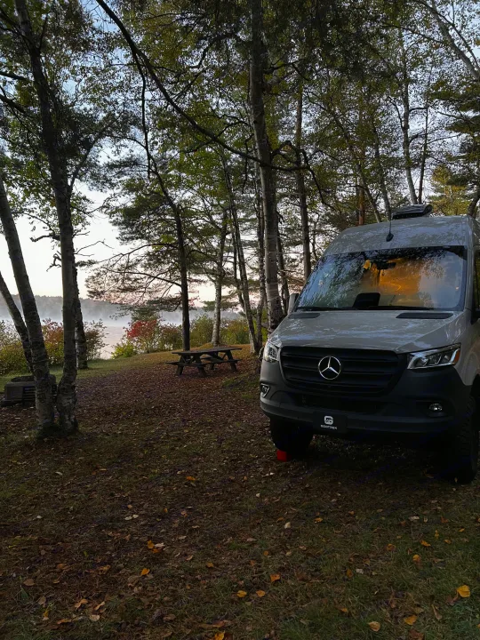 RV rental camper van parked by a misty lake, surrounded by autumn trees and a picnic table at a scenic campsite.. Mercedes Sprinter 2025