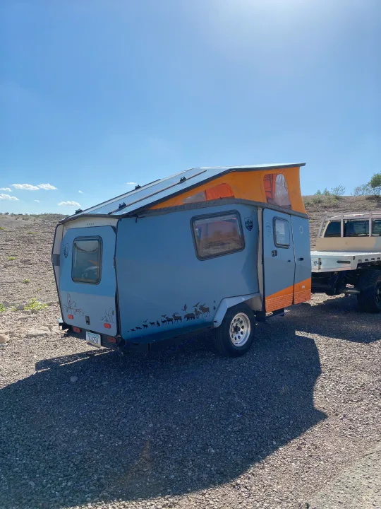 Unique gray & orange rv rental camper trailer with solar panels & pop-top, ready for outdoor adventure on rocky ground.. Cricket standard 2013
