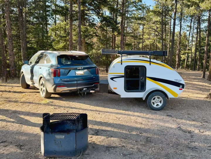 Blue SUV towing a white teardrop RV rental, parked at a wooded campsite. A fire pit is in the foreground, ready for camping.. Lil’ Guy RET 2006