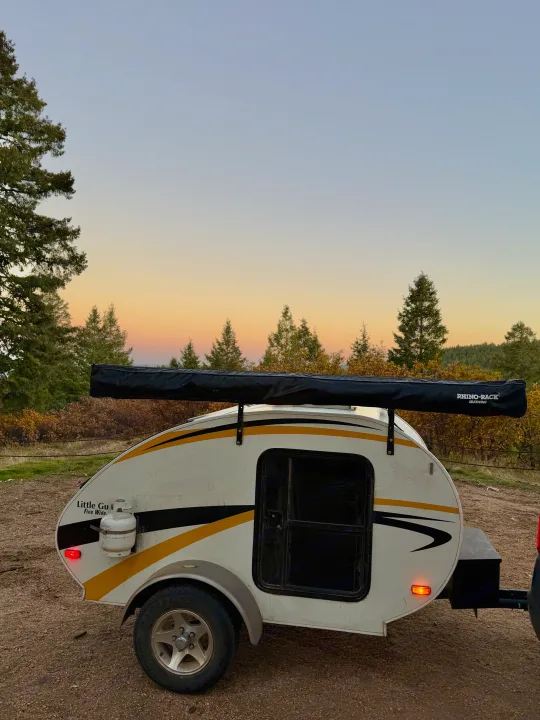 A white teardrop RV rental camper with a black awning under a golden sunset sky, surrounded by pine trees. Ready for camping.. Lil’ Guy RET 2006